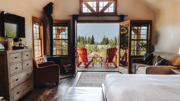 A bedroom with a large bed, a dresser, a TV, and a view of a patio with chairs and a table. - Heather Mountain Lodge (Golden)