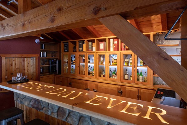 A bar area with wooden cabinets displaying liquor bottles, a stone countertop with 'BESTDAYEVER' signage, and a rustic wooden ceiling.