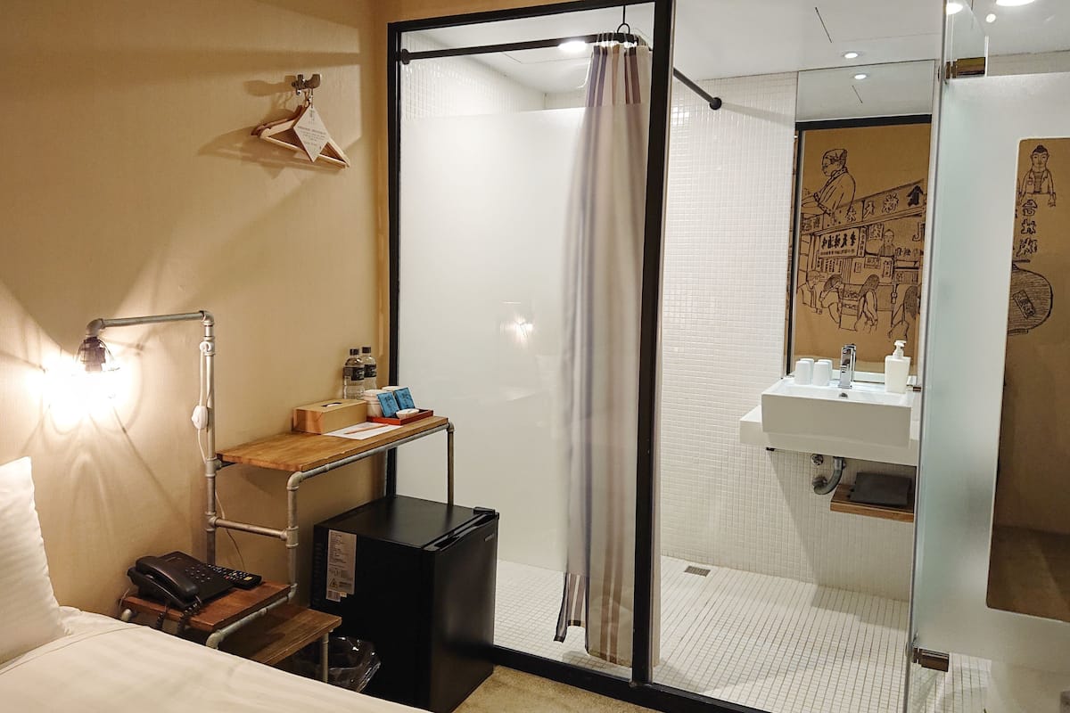 Une chambre d’hôtel comprenant un lit, un bureau avec une lampe, une petite table avec des articles de toilette, une poubelle noire et un espace douche doté d’une porte en verre.