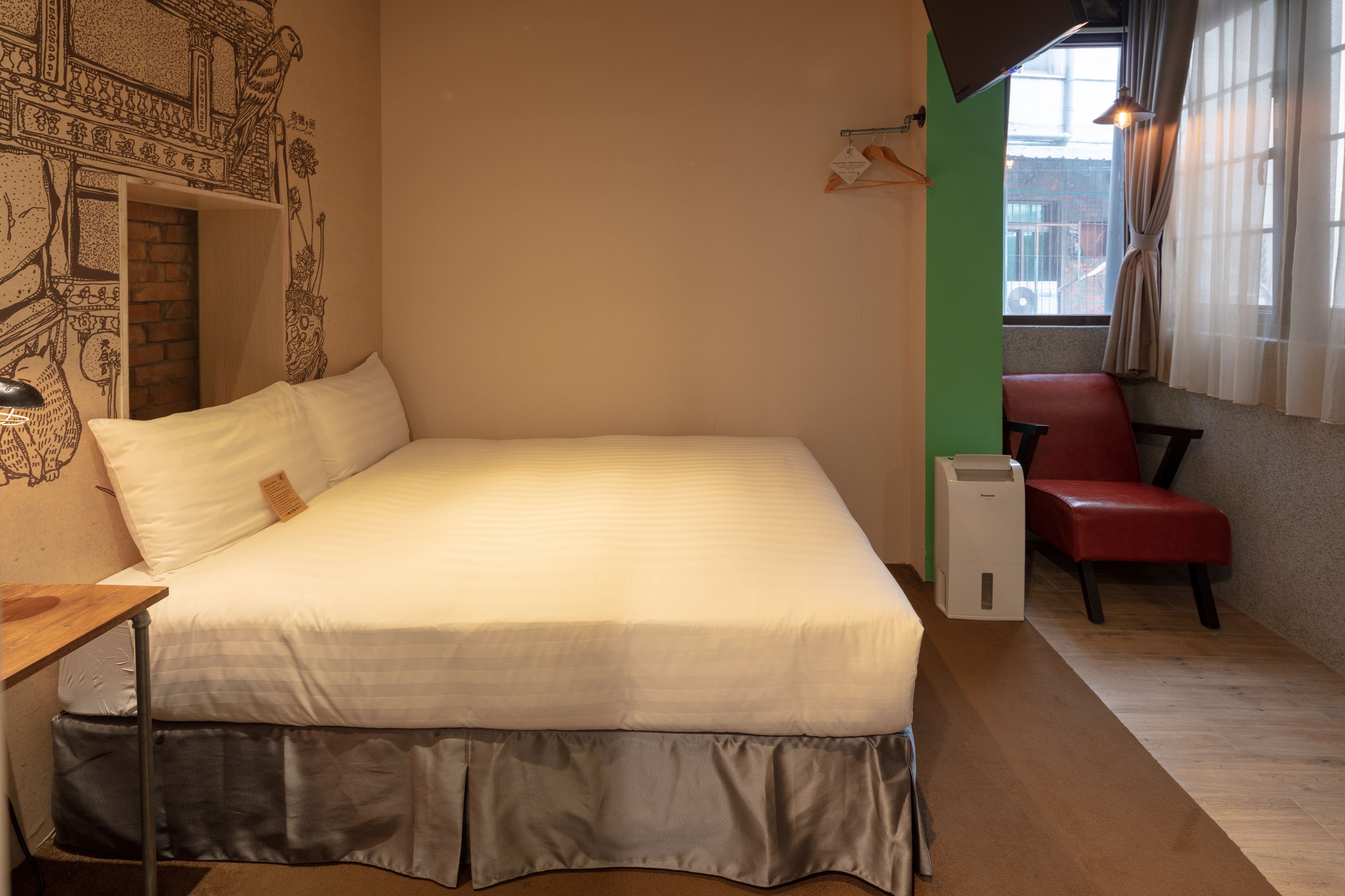 A neatly made bed with a white comforter, a red chair, a small table, and a wall with a decorative mural.