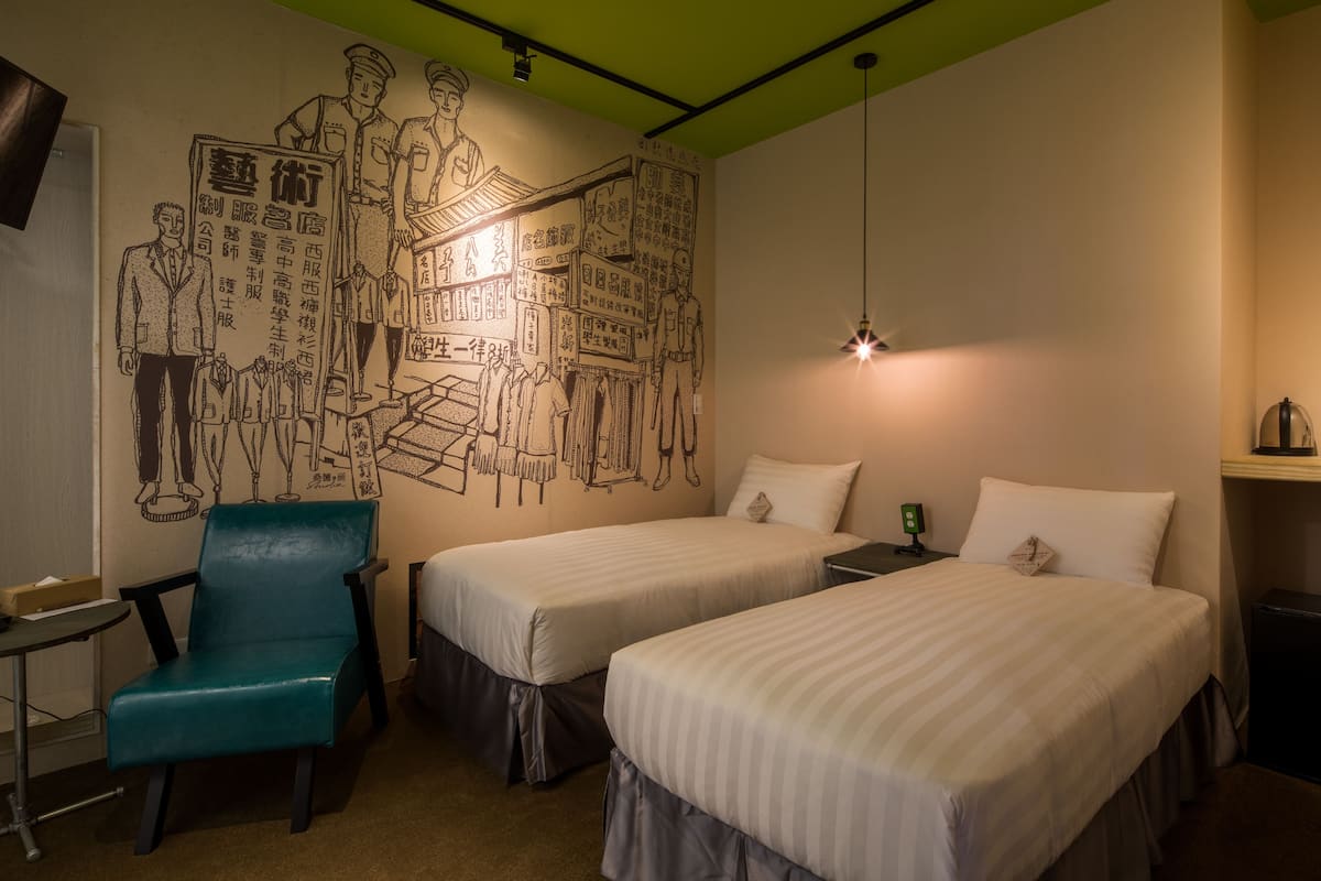 Une chambre d’hôtel avec deux lits, un fauteuil de couleur turquoise, un bureau et une fresque murale.
