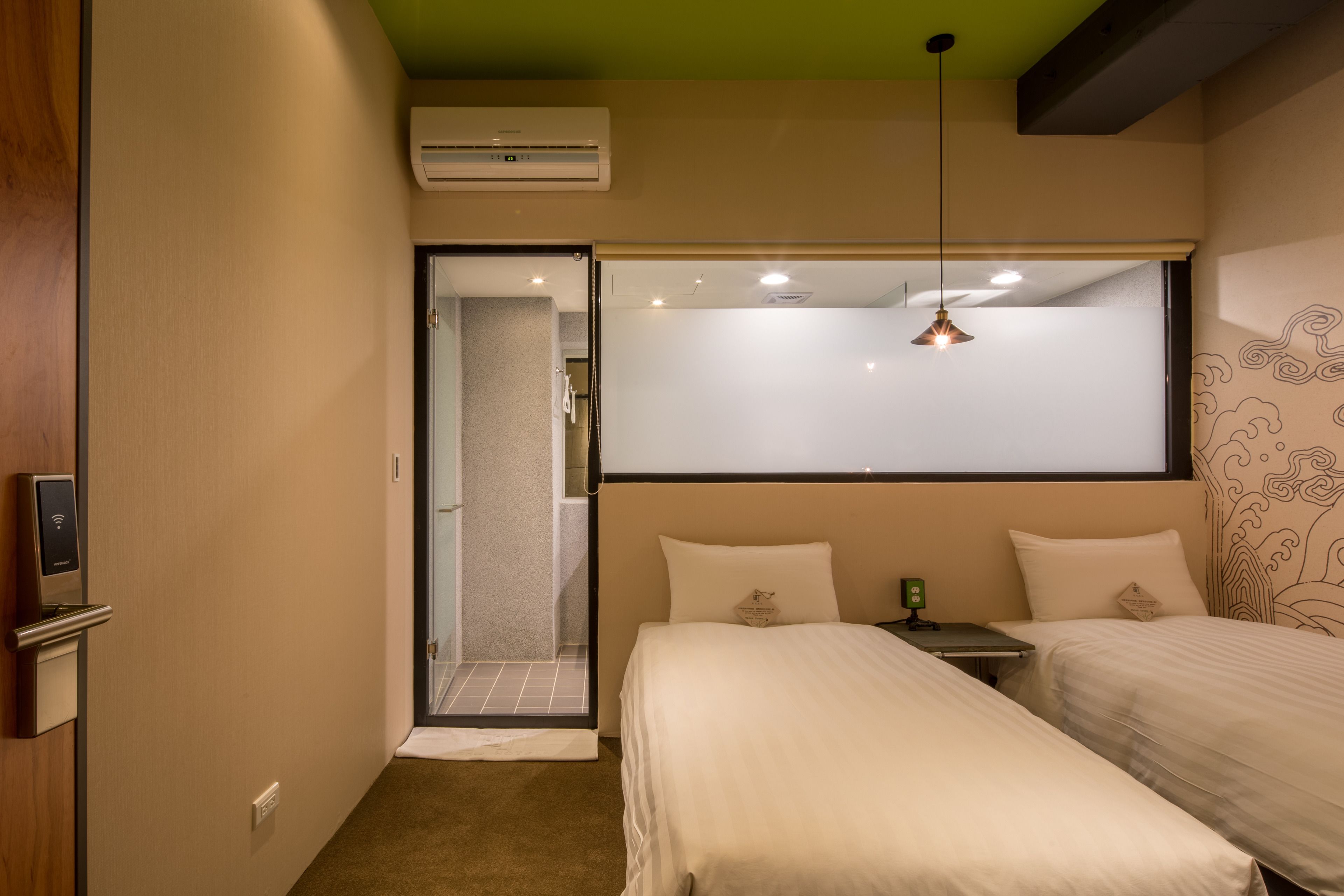 A hotel room with two beds, a small table, a wall-mounted air conditioner, and a ceiling light fixture.