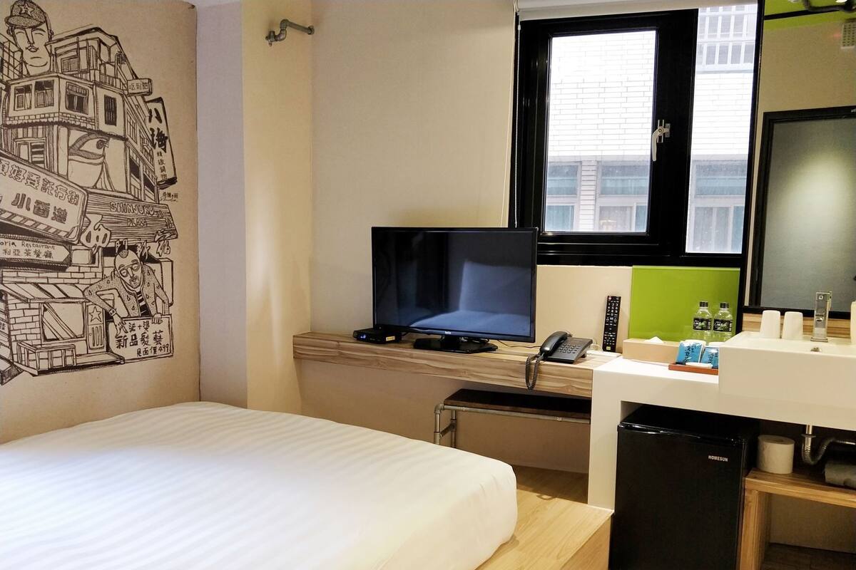 Une chambre d’hôtel équipée d’un lit, d’un bureau avec un ordinateur et un téléphone, d’un petit réfrigérateur et d’une fenêtre donnant sur des immeubles.