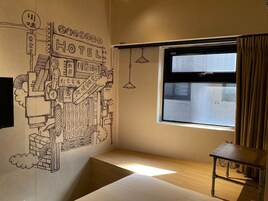 Een hotelkamer met een groot raam, een houten bankje en een muurschildering met diverse elementen die met een hotel te maken hebben.