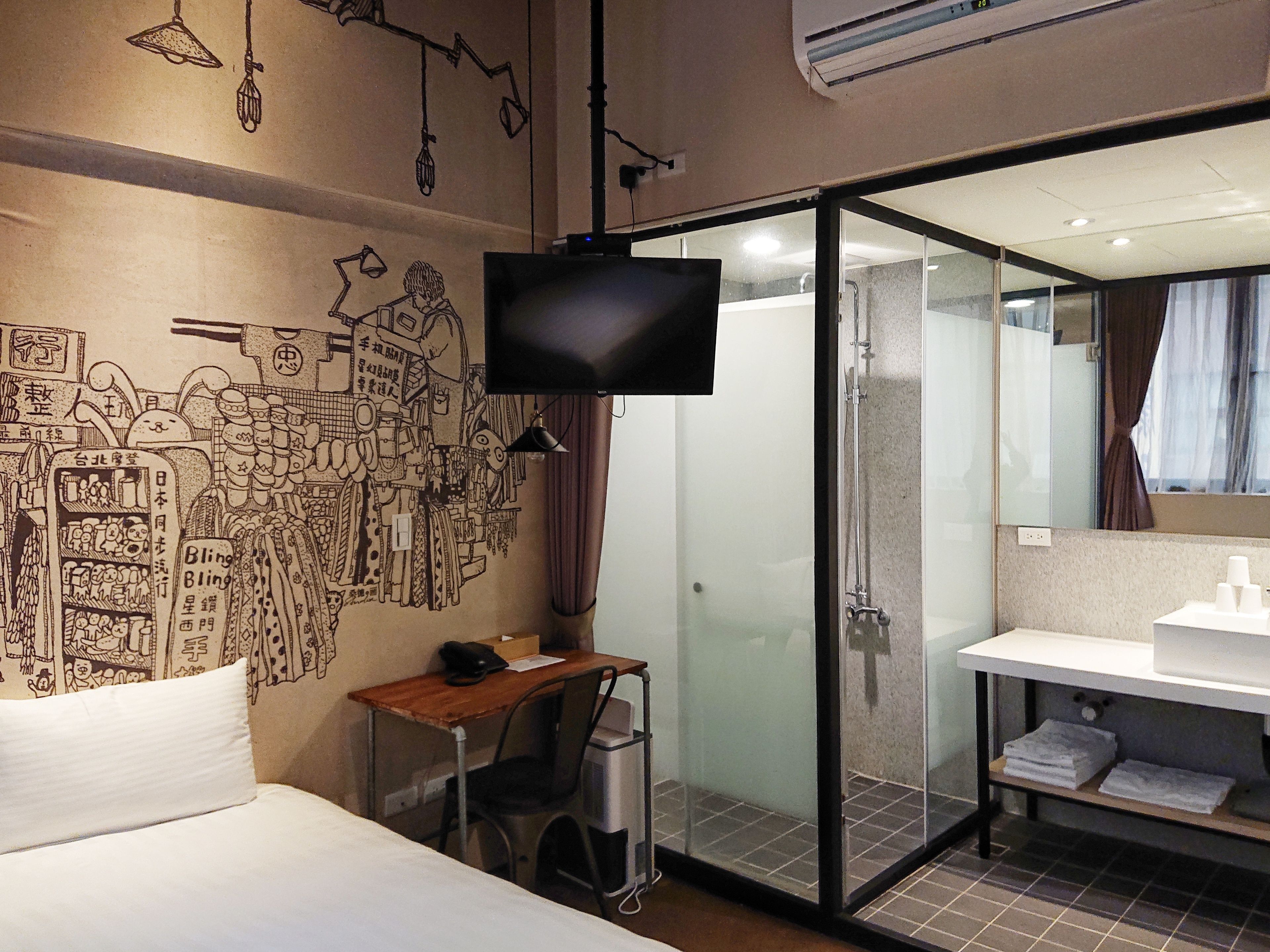A hotel room with a bed, a desk with a chair, a TV, a glass shower enclosure, and a bathroom with a sink and mirror.