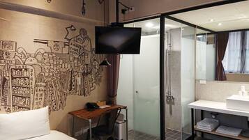 A hotel room with a bed, a desk with a chair, a TV, a glass shower enclosure, and a bathroom with a sink and mirror.