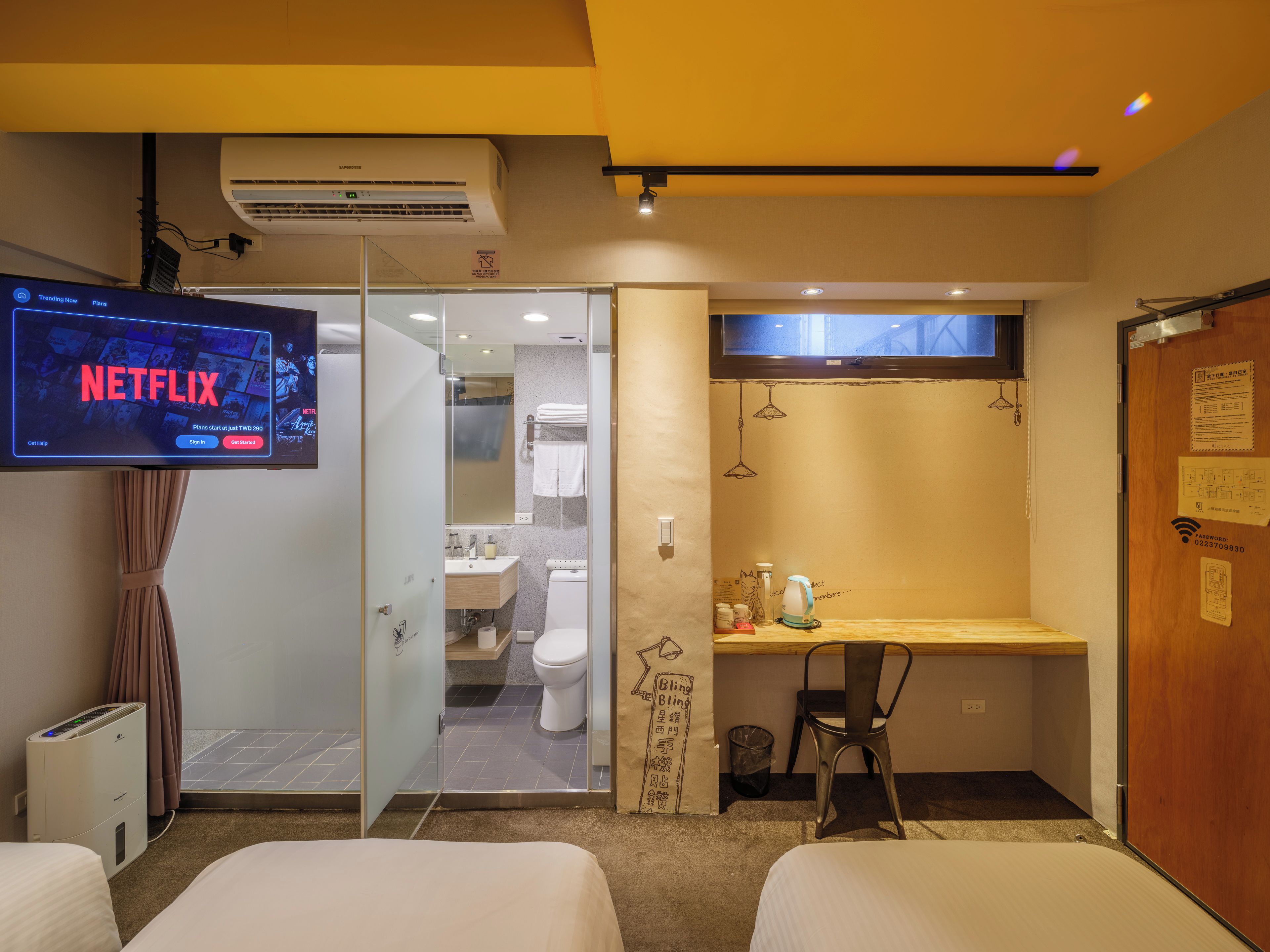 Quarto de hotel com televisão exibindo a interface da Netflix, banheiro com vaso sanitário e pia, e escrivaninha de madeira com cadeira.