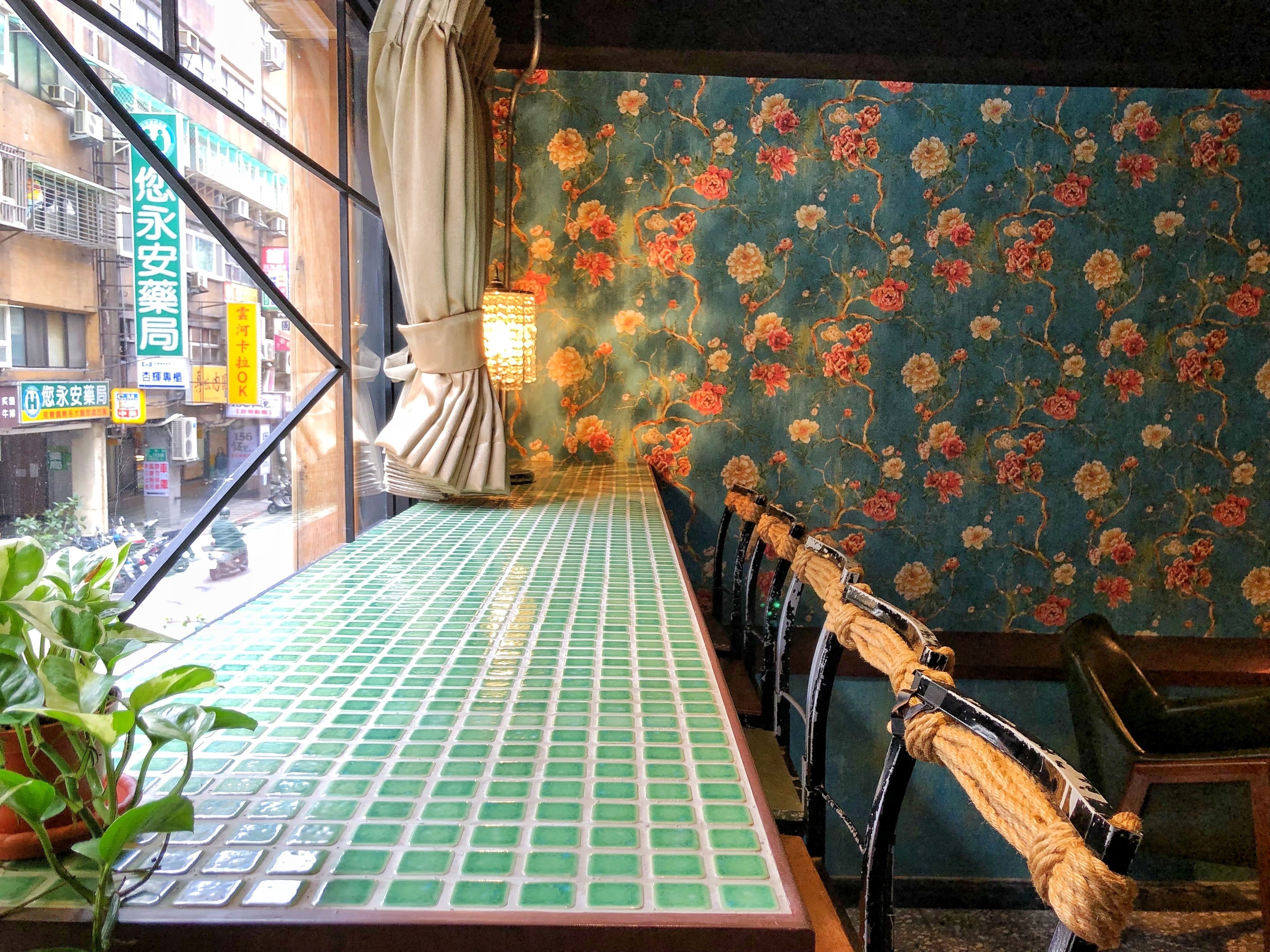 A room with a green tiled floor, a window with a view of the street, and a floral wallpaper.