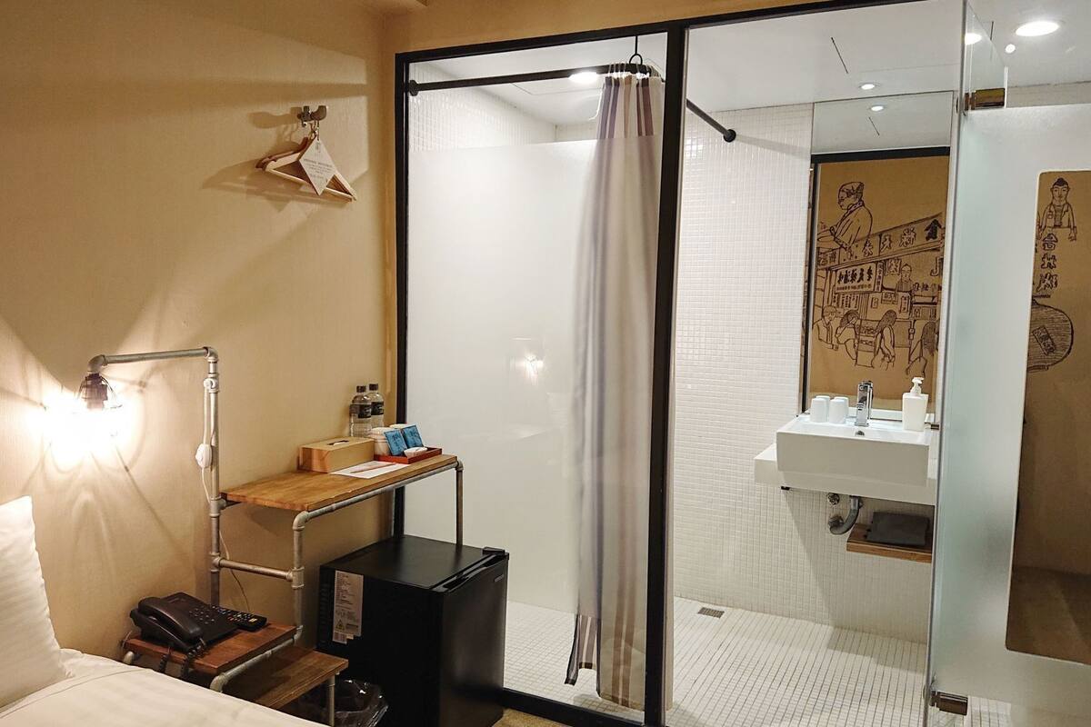 Une chambre d’hôtel comprenant un lit, une table de chevet avec une lampe, un petit bureau avec un téléphone et un boîtier noir, un espace douche avec un rideau, et une salle de bain avec un lavabo et un miroir.
