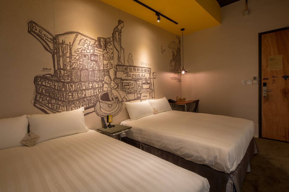 Une chambre d’hôtel avec deux lits, une fresque murale, une porte en bois et une petite table avec une lampe.