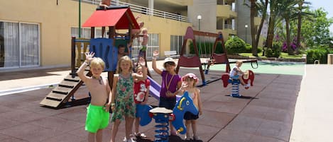 A playground with various play structures, including a slide and swings, in front of a building with balconies and palm trees.