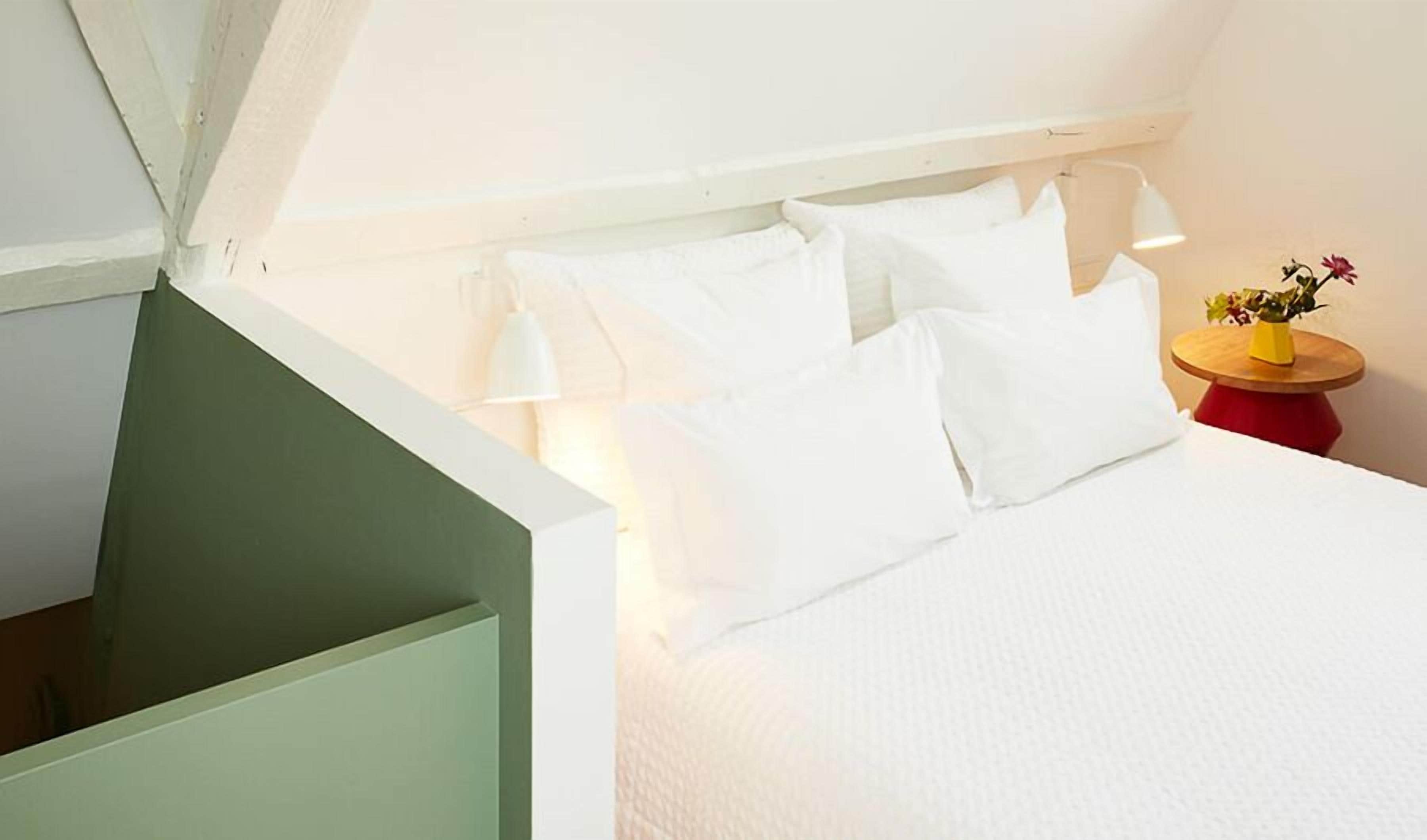 A bedroom with a white bed, white pillows, a small wooden side table with a yellow vase, and a red base.
