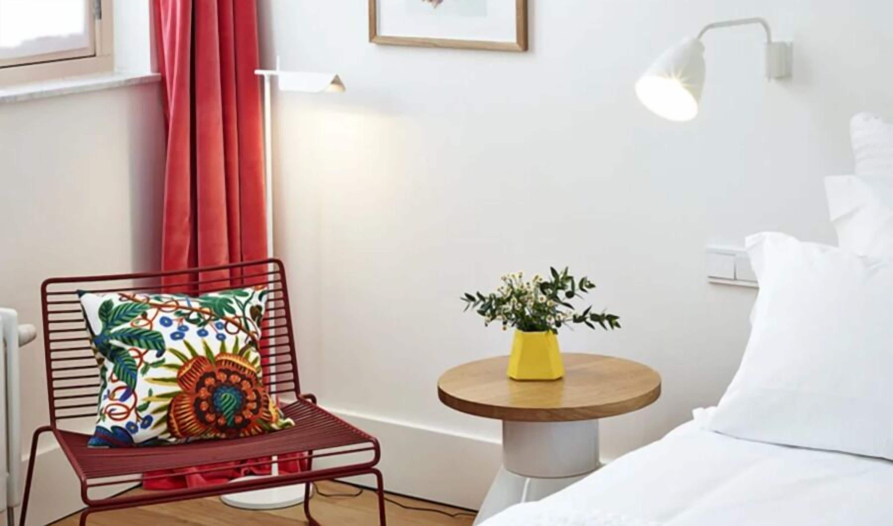 Ein Hotelzimmer mit einem Bett, einem roten Sessel mit bunten Kissen, einem kleinen runden Tisch mit einer gelben Vase und einem Fenster mit roten Vorhängen.