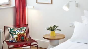 Ein Hotelzimmer mit einem Bett, einem roten Sessel mit bunten Kissen, einem kleinen runden Tisch mit einer gelben Vase und einem Fenster mit roten Vorhängen.