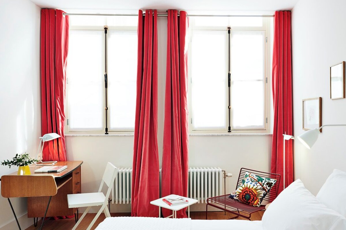 A hotel room with a bed, a desk with a chair, a radiator, and a window with red curtains.