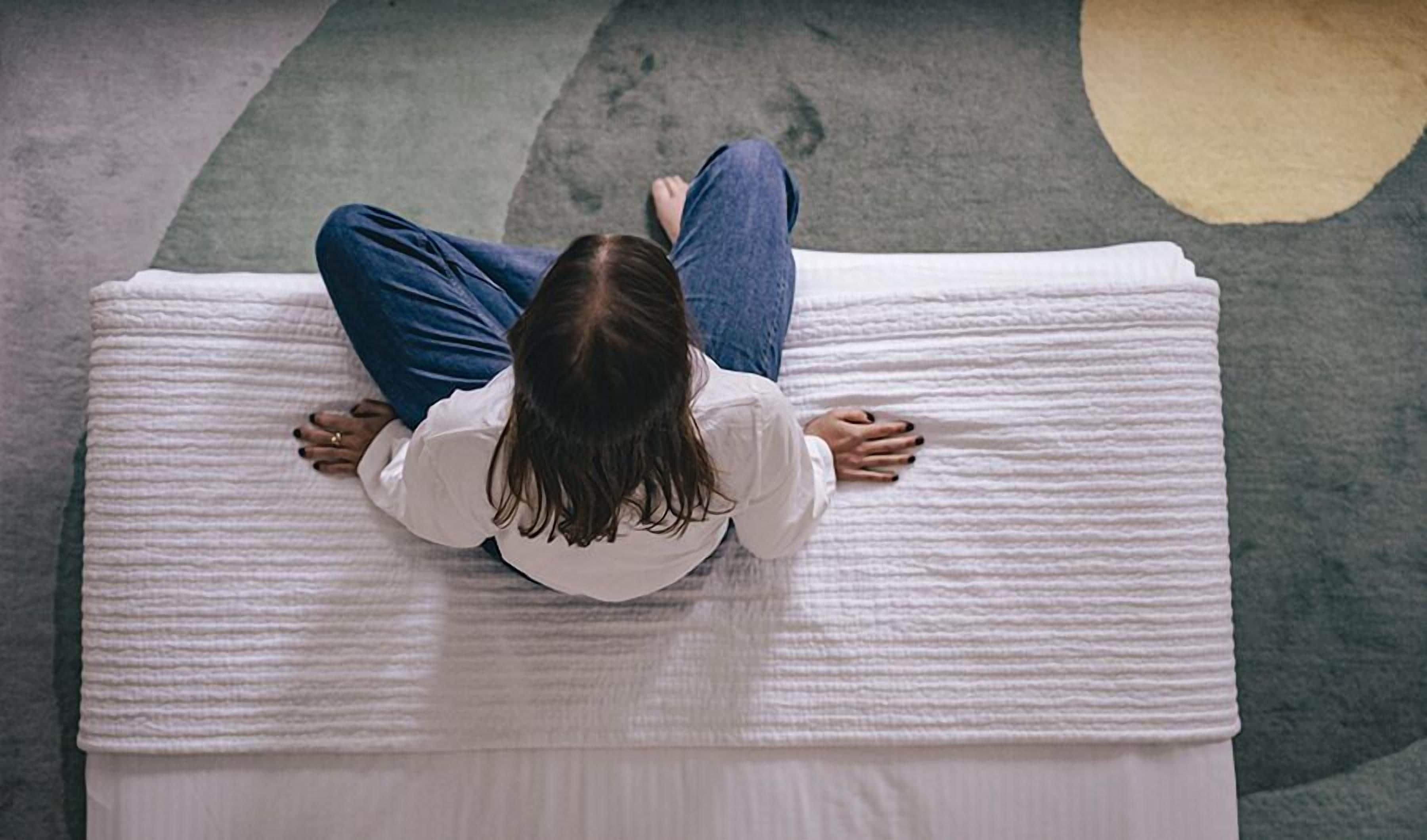 Una persona acostada sobre un colchón blanco en una habitación con alfombra gris y un tapete circular.