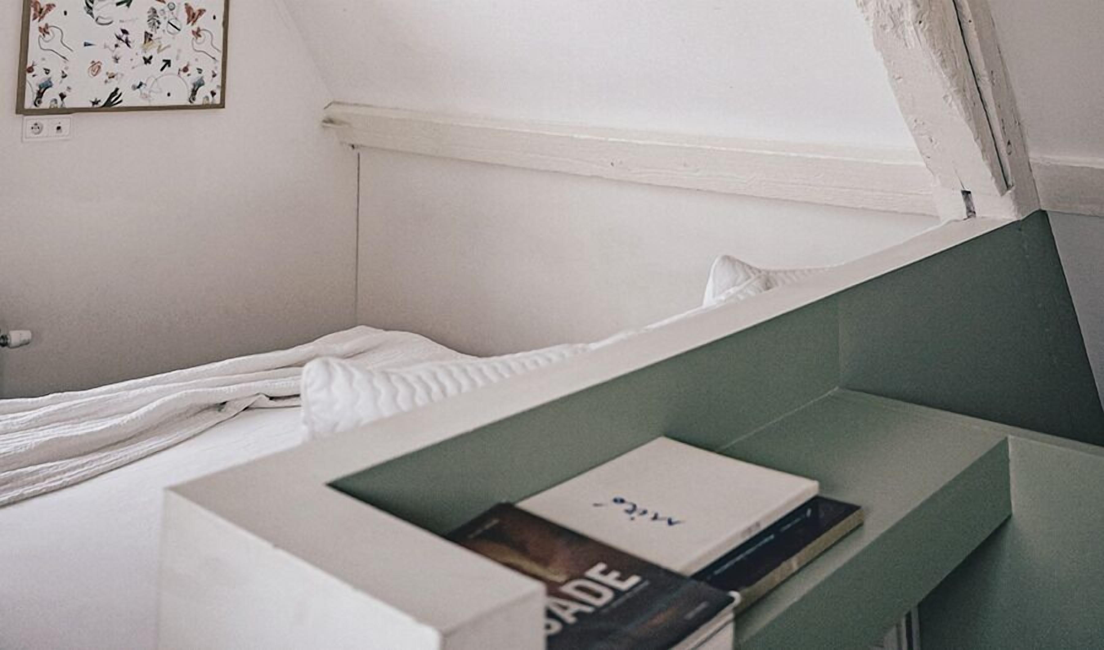 Una cama bien hecha con edredón blanco y almohadas, una mesita de noche con un libro, y una pequeña obra de arte en la pared encima de la cama.