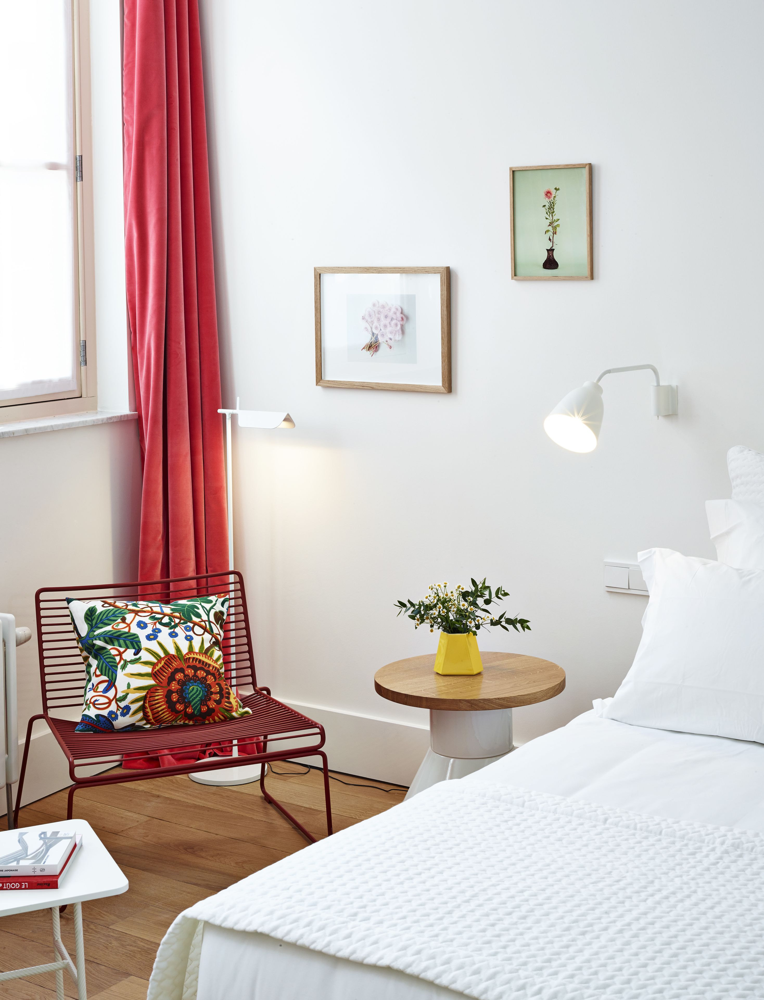 A hotel room with a bed, a chair with a colorful cushion, a small table with a yellow vase, and a wall-mounted lamp.