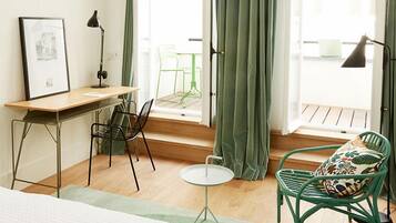 A small, bright room with a wooden desk, two chairs, a small table, and a bed.