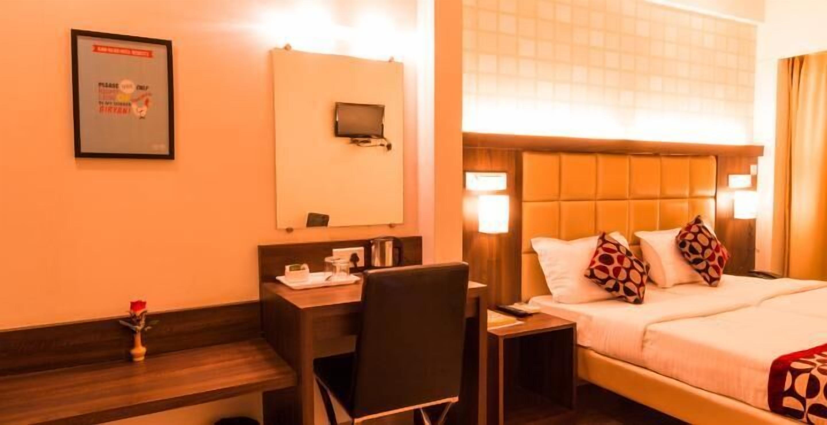 Quarto de hotel com cama, escrivaninha, cadeira, mesa pequena, mesa de cabeceira, televisão e um letreiro emoldurado na parede.