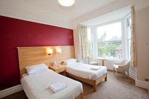 A hotel room with two beds, a wooden headboard, a small table, a chair, and a window with curtains.