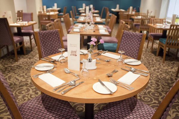 A dining area with round tables set for four, each with white plates, silverware, and glassware. There are purple upholstered chairs and a menu stand with a floral centerpiece. - Wheatlands Lodge Hotel (York)