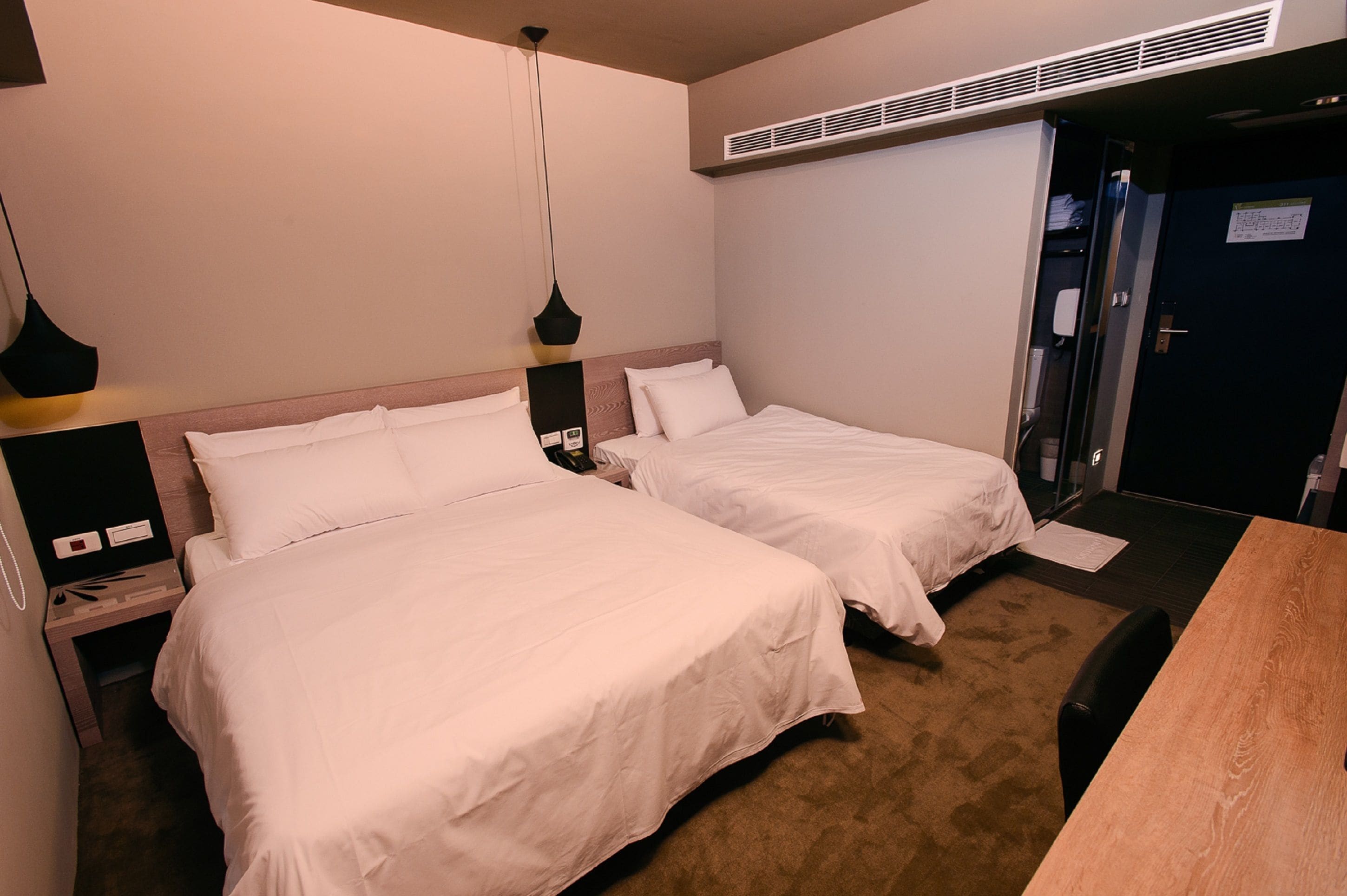 Quarto de hotel com duas camas, uma mesa de madeira e uma escrivaninha com cadeira.