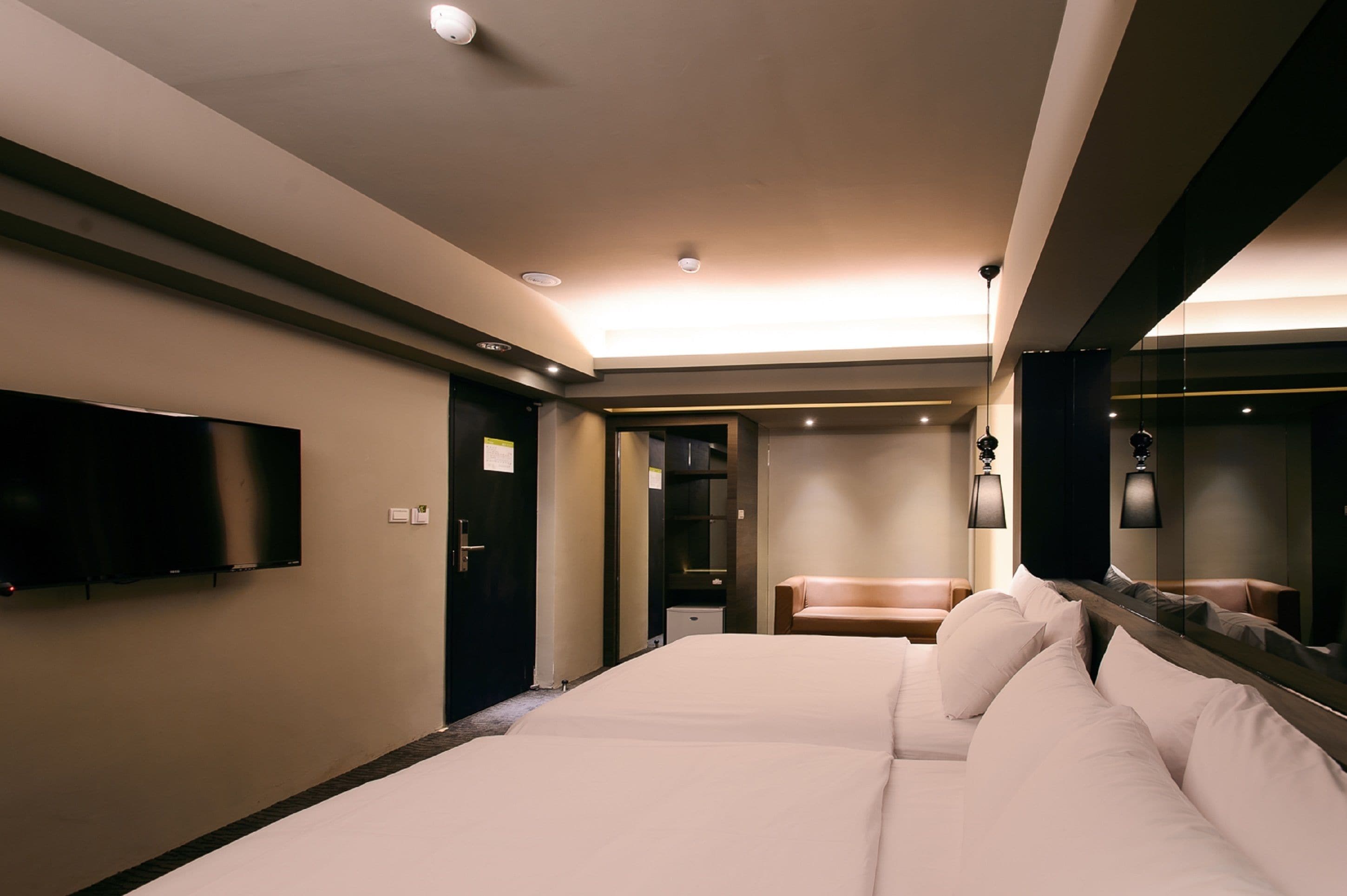 A modern hotel room with a large bed, a flat-screen TV mounted on the wall, a bedside table, and a mirror.