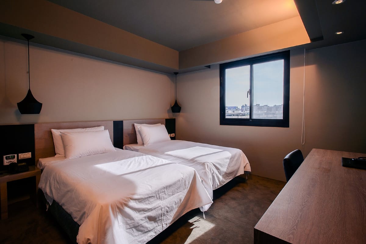Ein Hotelzimmer mit zwei Betten, einem Schreibtisch und einem Fenster mit Blick auf die Stadt.