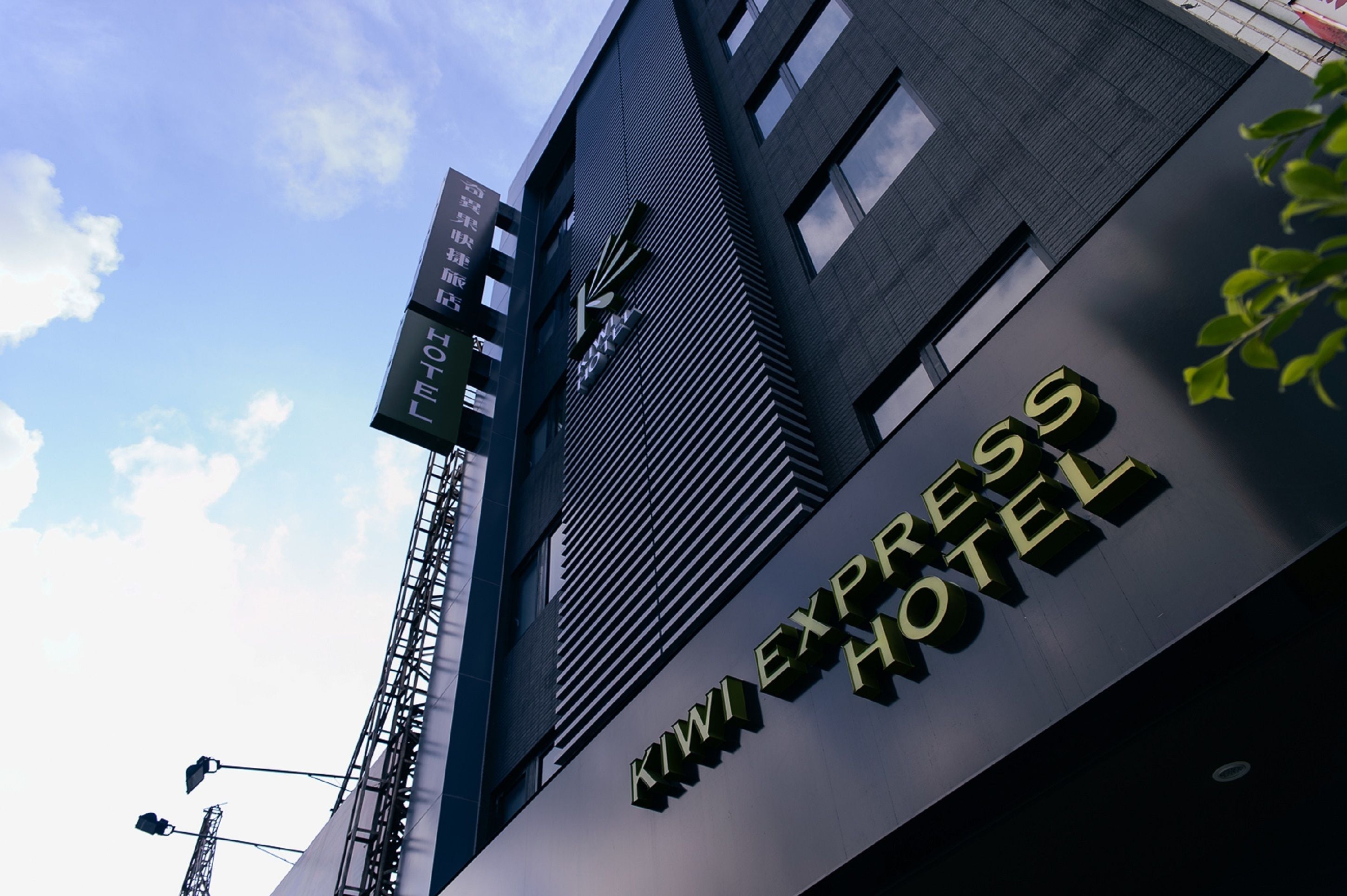 Exterior del Hotel Kiwi Express, con un diseño moderno, destacando un letrero llamativo y un cielo despejado al fondo.