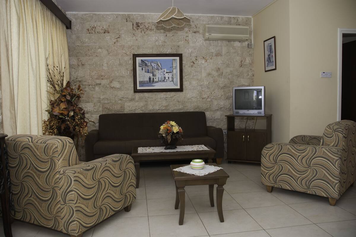 Una sala de estar con sofá, dos sillones, una mesita de centro, televisor sobre un mueble bajo y un cuadro en la pared.