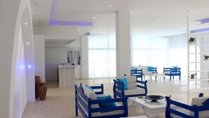 Uma sala de estar moderna com móveis azuis e brancos, grandes janelas e uma cozinha ao fundo.
