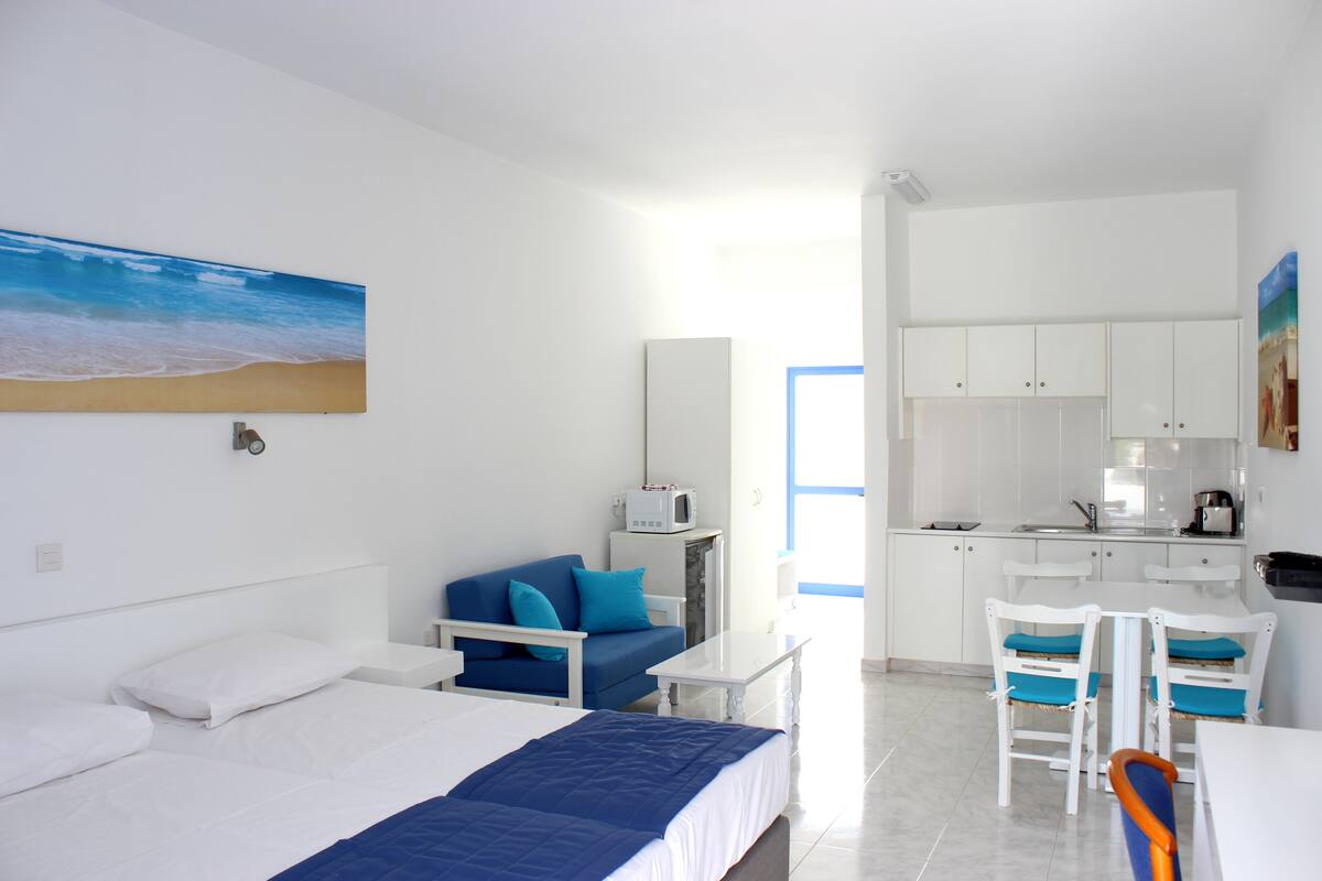 Une chambre d’hôtel équipée d’un lit, d’un bureau, d’une chaise, d’une kitchenette et d’un tableau représentant une plage.