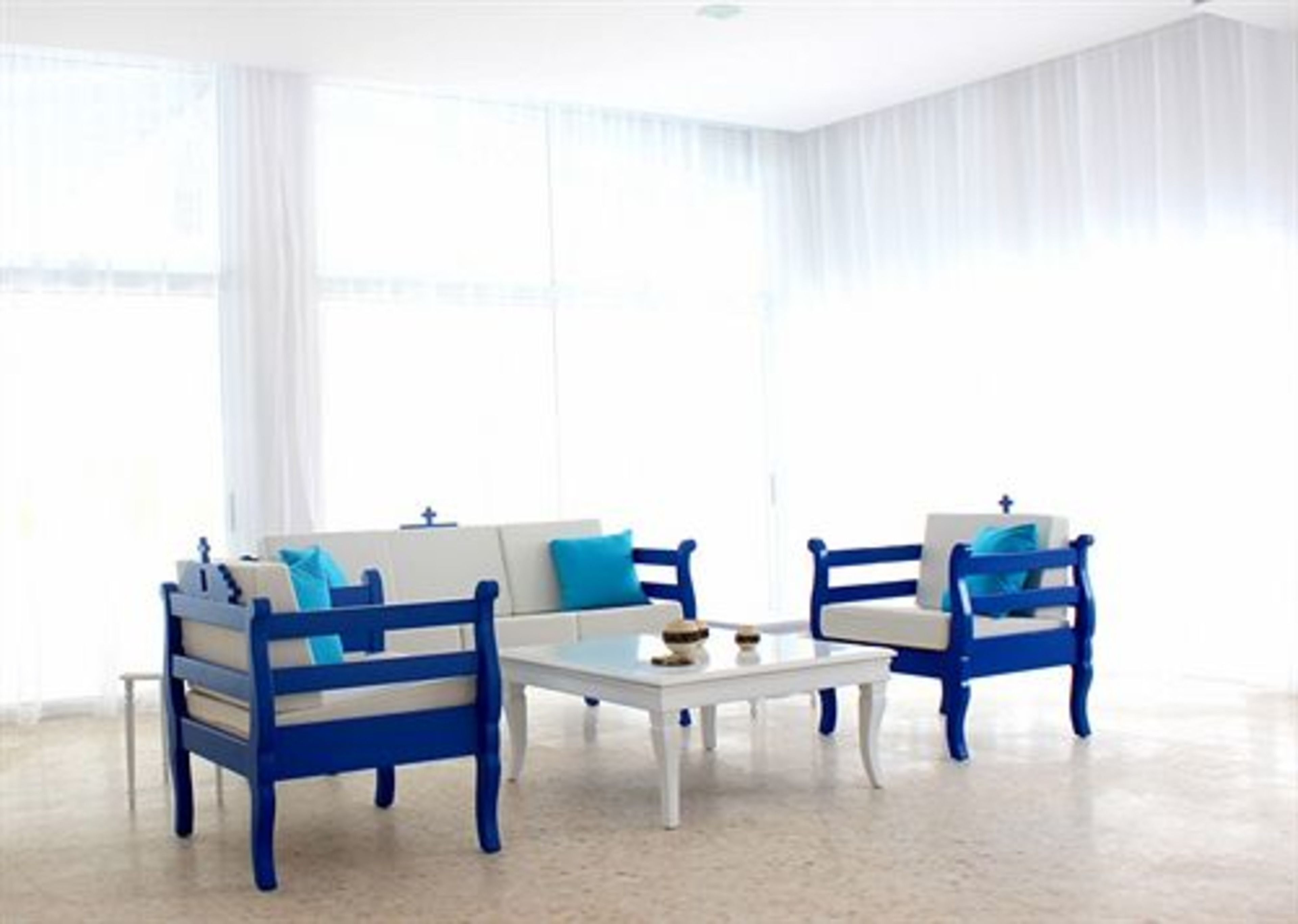 A room with a white sofa and two blue armchairs, a small white table, and sheer white curtains.