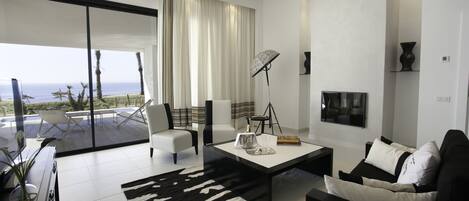 A modern living room with a large sliding glass door leading to a balcony with a view of the sea. There is a black and white patterned rug, a black coffee table, and a white sofa with black cushions.
