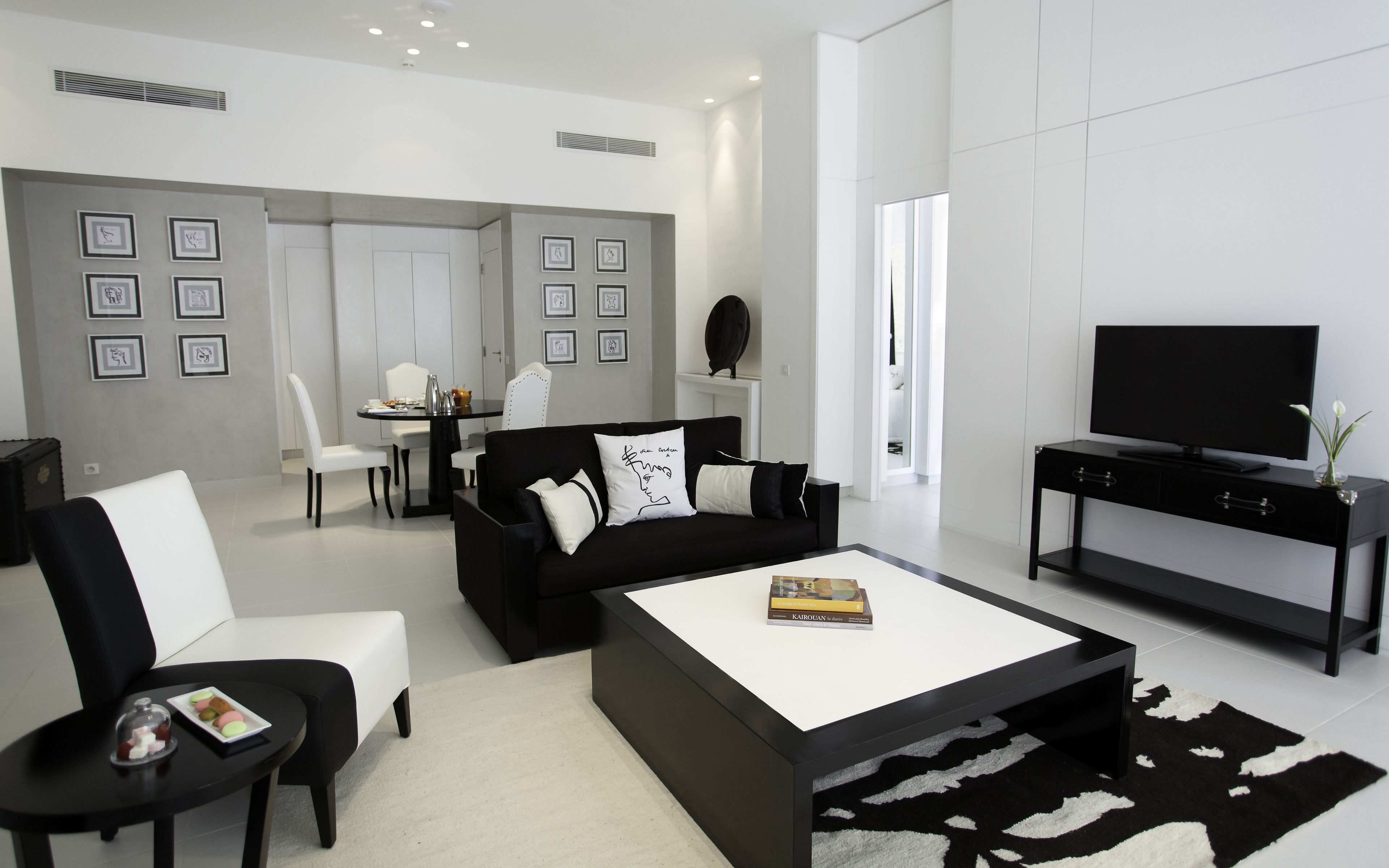 A modern living room with a black sofa, white armchairs, a coffee table, and a TV on a black stand. There is a dining area with a white table and chairs in the background.