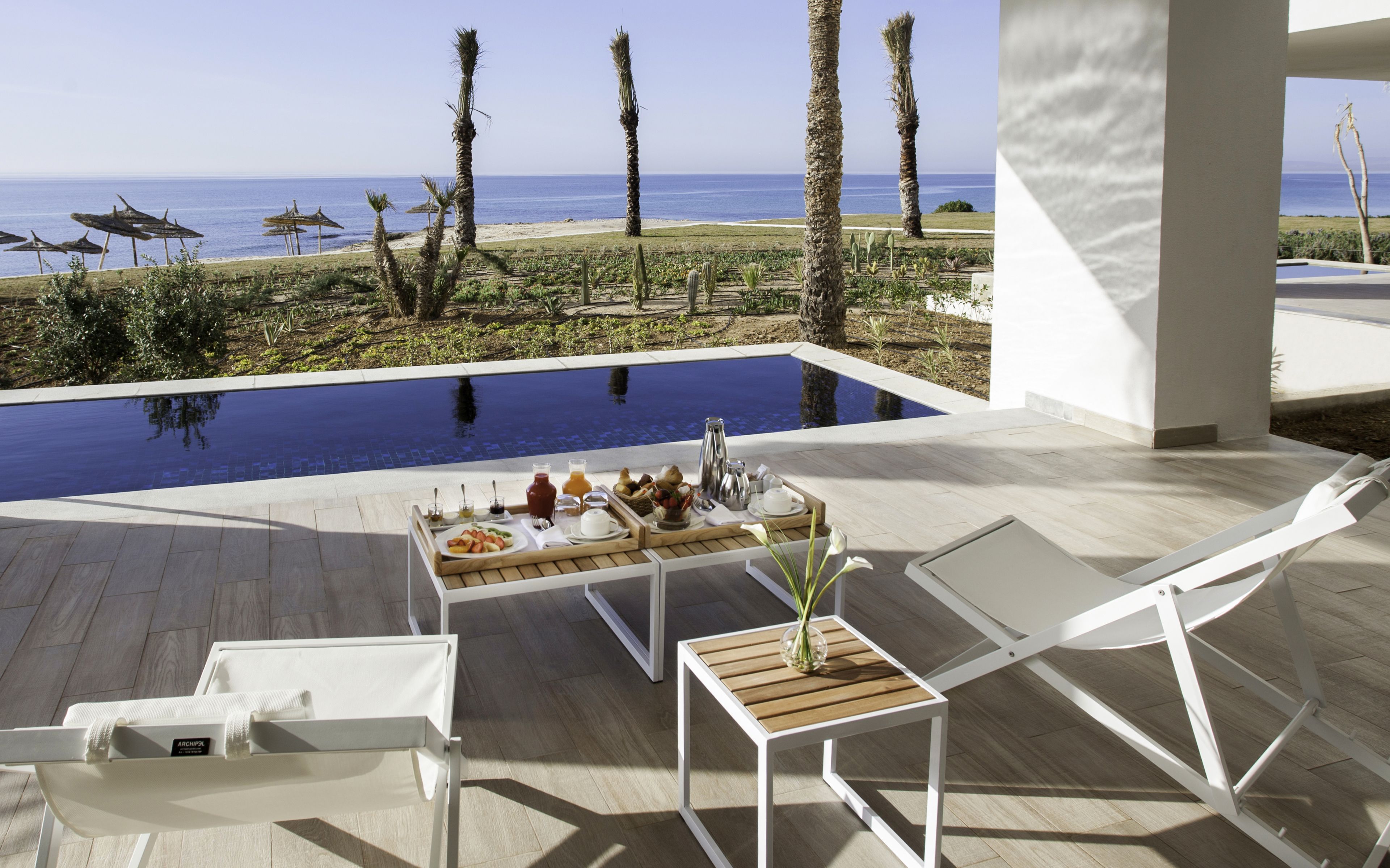 Ein Poolbereich mit Liegestühlen, einem Tisch mit Frühstücksangeboten und Blick auf den Ozean.