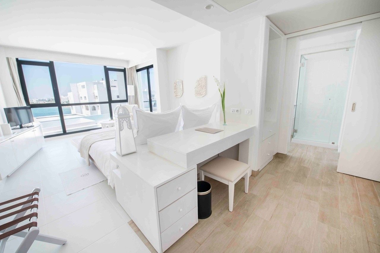 Quarto moderno com decoração em tons de branco, uma janela grande, cama, escrivaninha e um banquinho.