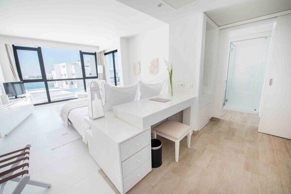 Un dormitorio moderno con decoración en tonos blancos, una ventana grande, una cama, un escritorio y un banco.