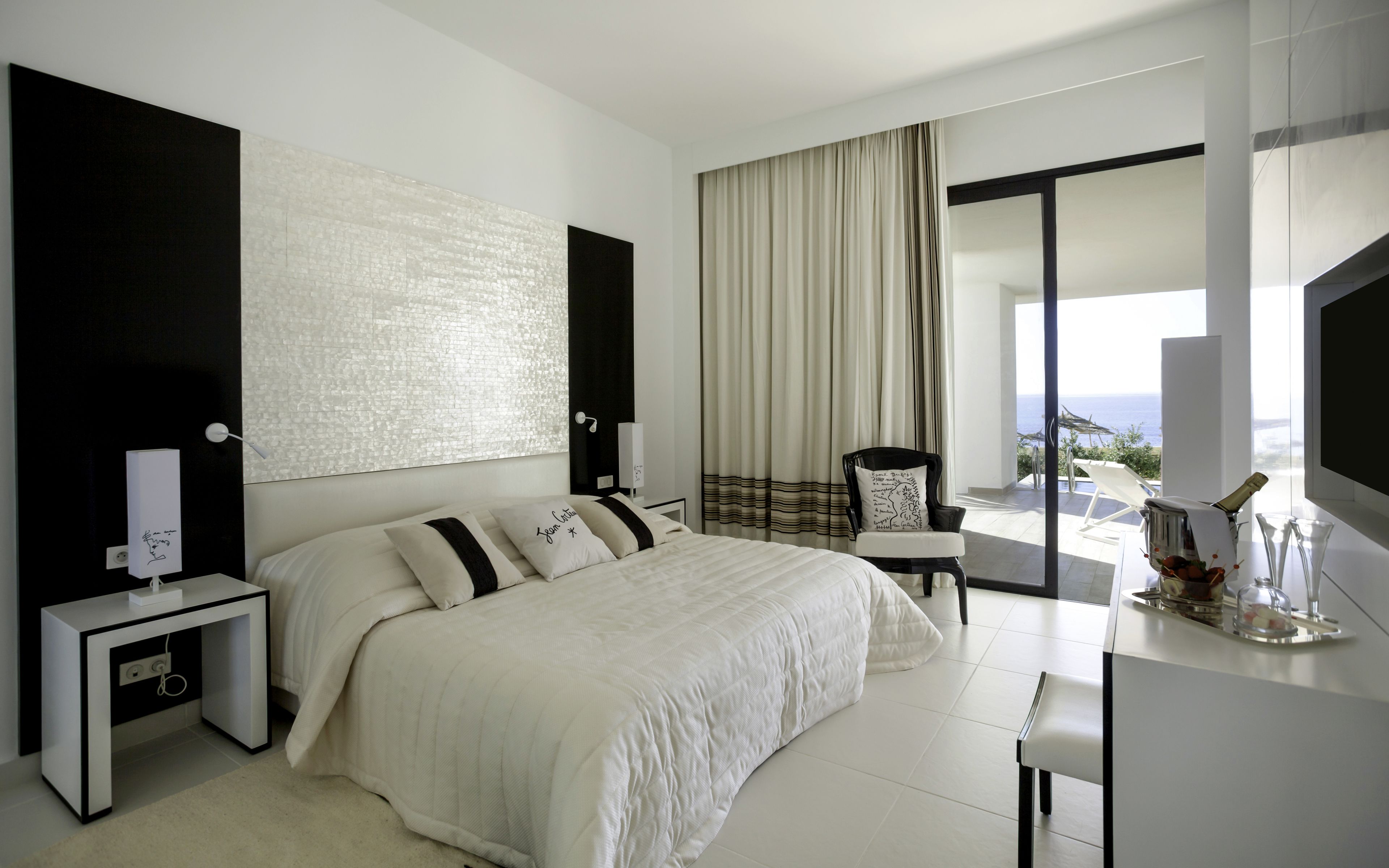 A modern bedroom with a large bed, a desk, a chair, and a view of the ocean.
