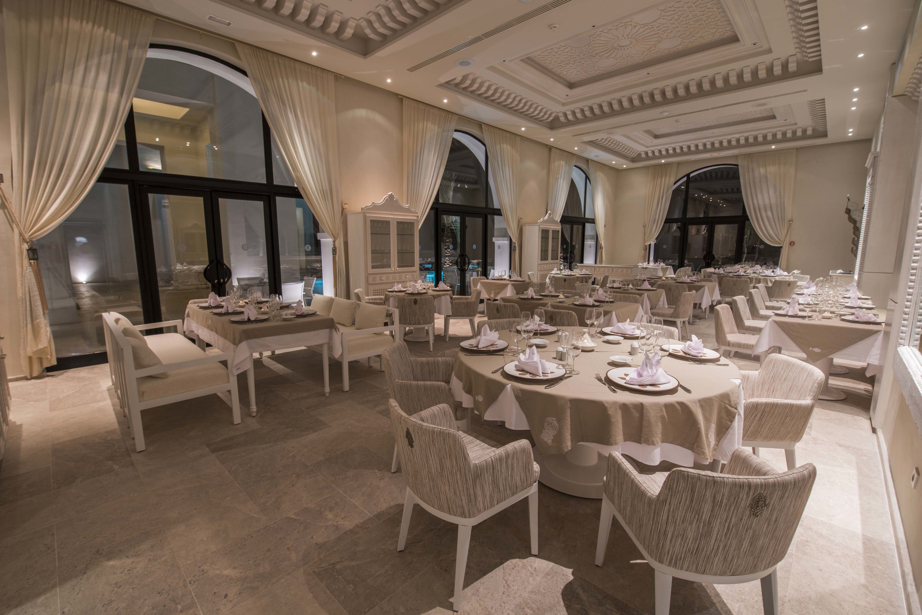 A formal dining room with round tables, white chairs, and chandeliers.