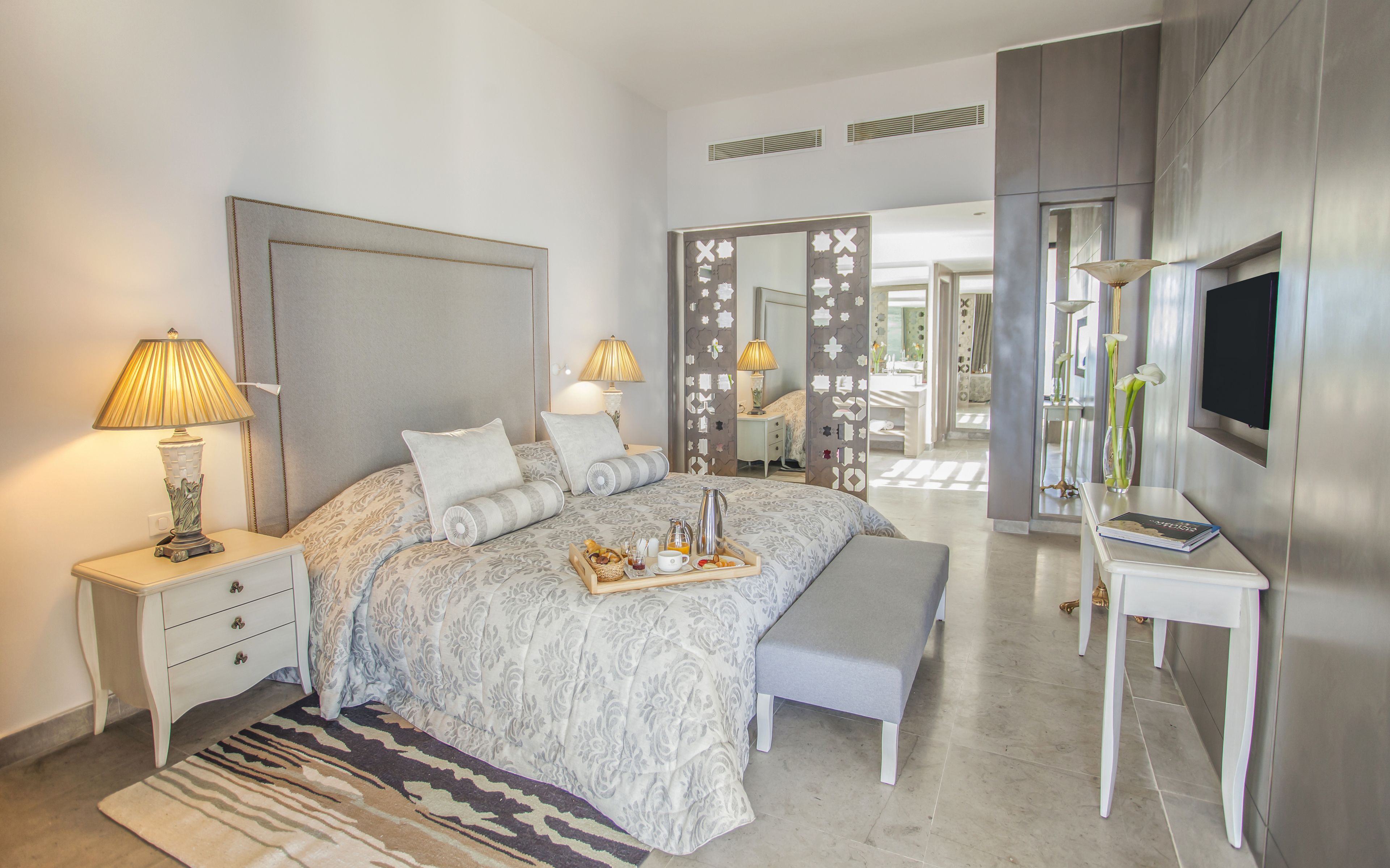 A modern bedroom with a large bed, bedside lamps, a bedside table, a bench, a TV, and a mirror.