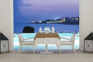 Una mesa de comedor con sillas blancas y mantel, con vistas a una piscina y al paisaje urbano al anochecer.