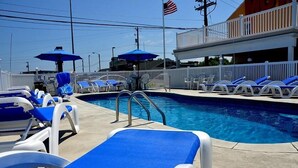 A swimming pool with blue lounge chairs and umbrellas, a white fence, and a building with a red awning in the background. - The Aventis Inn (Somers Point)