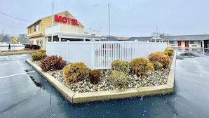 A motel with a prominent 'MOTEL' sign, a fenced parking area, and a variety of shrubs and plants. - The Aventis Inn (Somers Point)