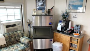 A compact hotel room with a kitchenette, a sofa, a trash bin, and a small wooden cabinet with books. - The Aventis Inn (Somers Point)