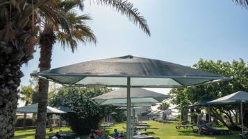 Un espacio al aire libre soleado con palmeras, mesas de picnic y sillones de descanso bajo un gran paraguas.