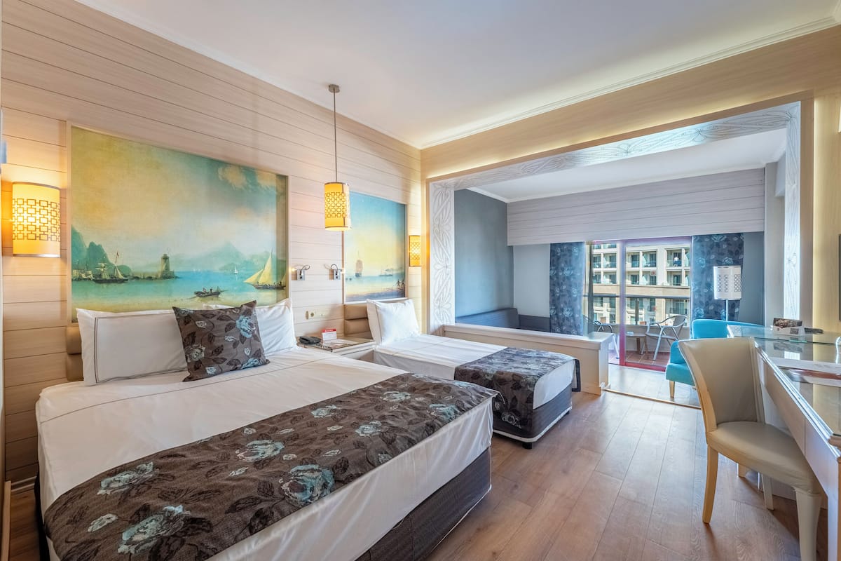 A hotel room with two beds, a desk, a chair, and a view of the sea through a large window.