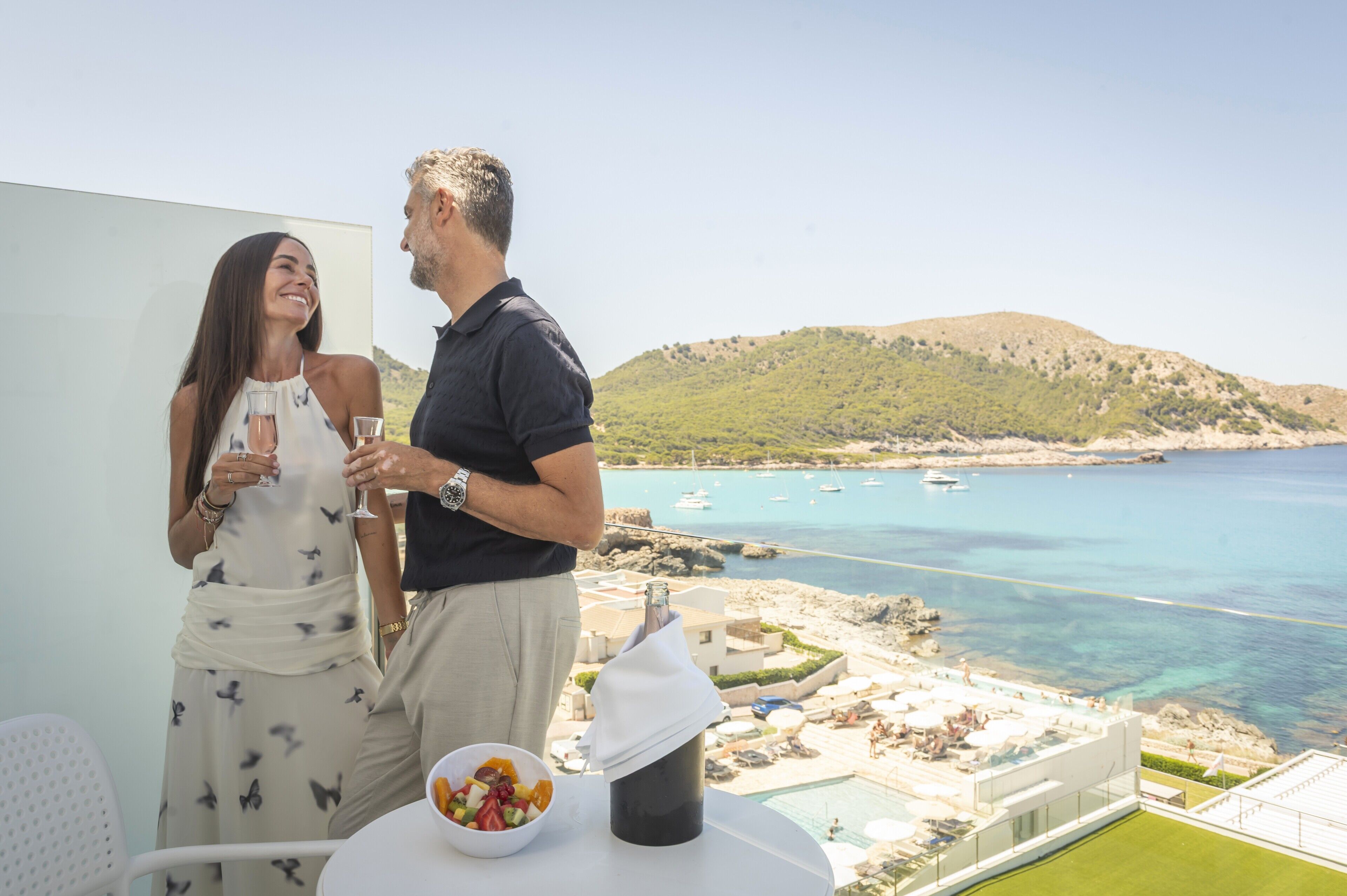 A couple standing by a balcony with a view of a coastal area, a pool, and a bottle of champagne.
