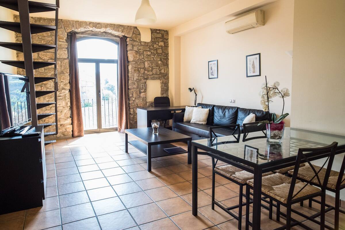 A spacious living room with a stone wall, a black leather sofa, a glass coffee table, and a dining area with wicker chairs.
